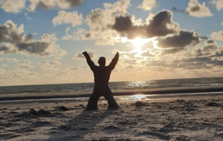 Ein jubelndes Männchen, geformt aus Sand, steht im Sonnenuntergang am Strand.